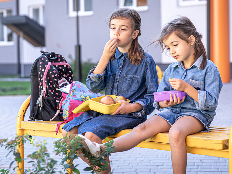 Little school girls sitting on bench in school yard and eating from lunch boxes.