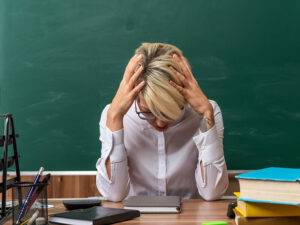 stressed young blonde female teacher wearing glasses sitting at desk with school tools in classroom keeping hands on head